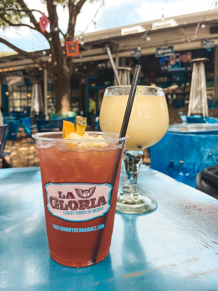 Two drinks sitting on a blue table outside at a Mexican restaurant in San Antonio. One is a red sangria in a disposable cup with a straw that says "La Gloria" on the front. The second, behind it, is a frozen margarita with a pale yellow color in a tall glass.
