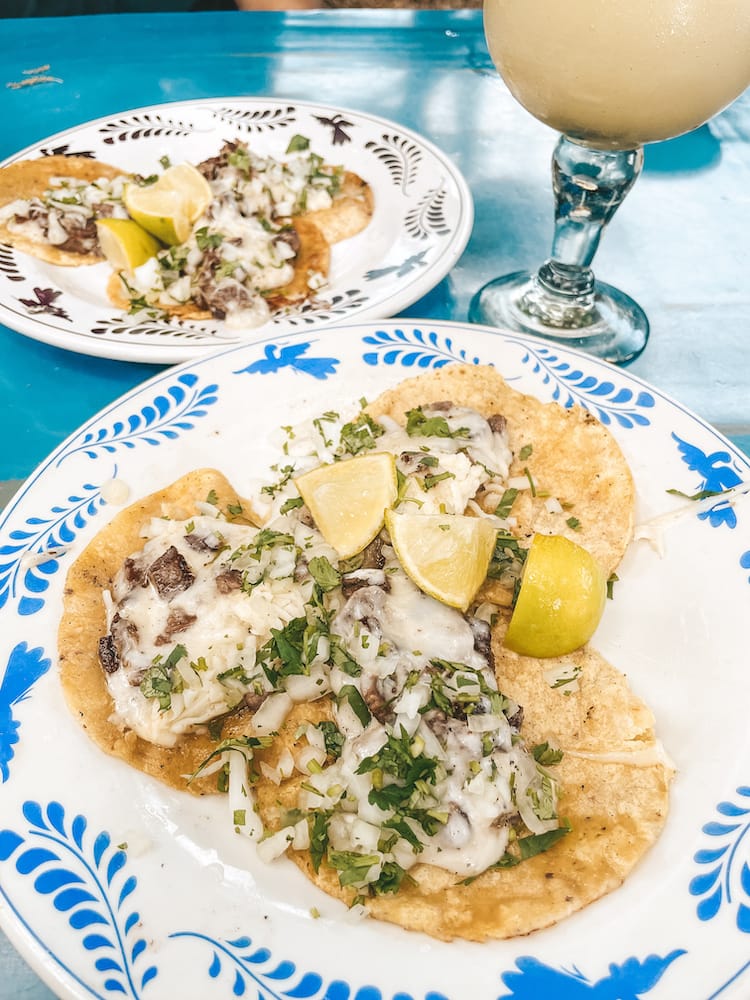 Two plates of beef, cheese, and cilantro tacos sitting on a blue table with a frozen margarita. Each plate has a blue, Spanish-style design on the outer rim that matches the table.