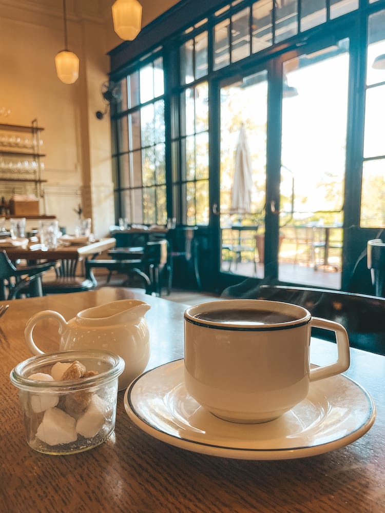 A cup of steaming coffee sitting on a saucer with a small bowl of sugar cubes and a small pitcher of cream sitting next to it.