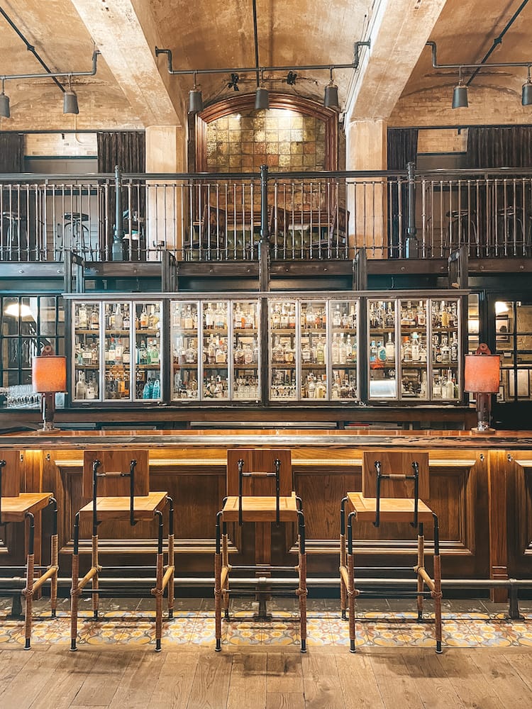 A historic industrial bar with wooden chairs, metal railing above, and several locked glass cabinets filled with alcohol.