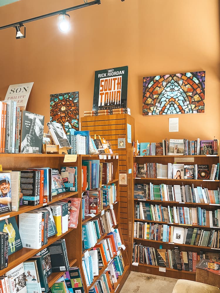 Two brown wooden bookshelves in a bookstore displaying several books in front of orange walls displaying colorful artwork.
