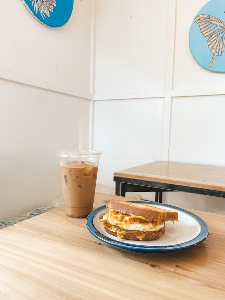 An iced coffee and a plate with a breakfast sandwich on it sitting on a wooden table in a white porch room