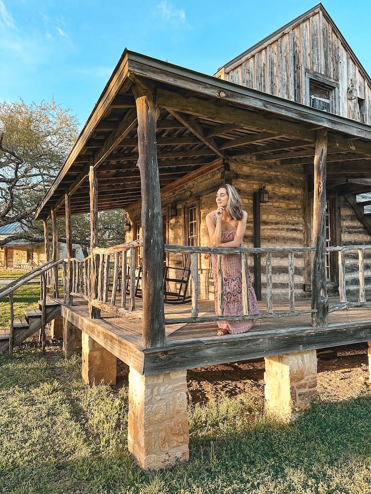 A woman in a pink floral dress standing on a wrap-around porch of a wooden log cabin.