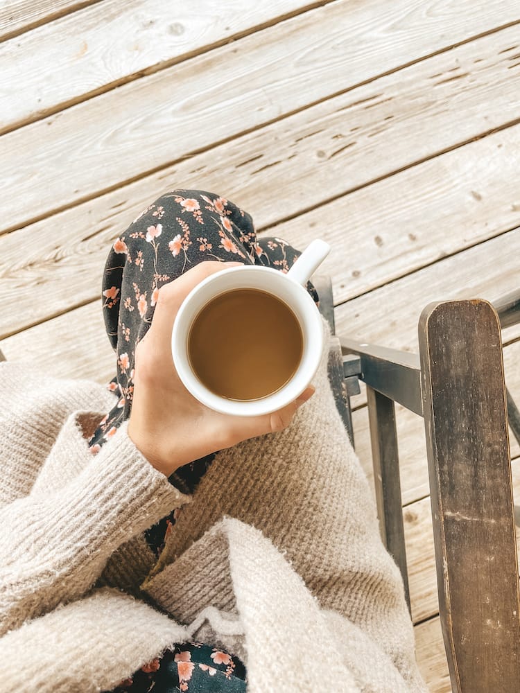 A cup of coffee in a while mug held by a woman in a sweater and pantsuit on a wooden porch