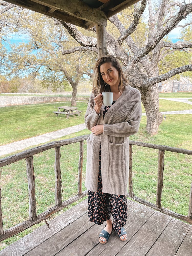 A woman standing in a sweater and pantsuit on a porch with trees in the background.