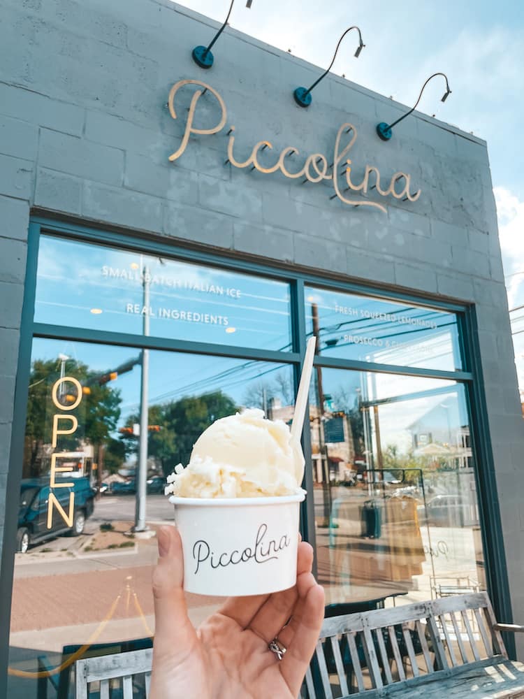 A scoop of pina colada Italian shave ice in front of a black building with "Piccolina," the name of the restaurant, in gold cursive letters