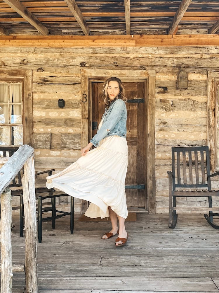 A woman in a white dress and denim jacket twirling around on a wooden front porch