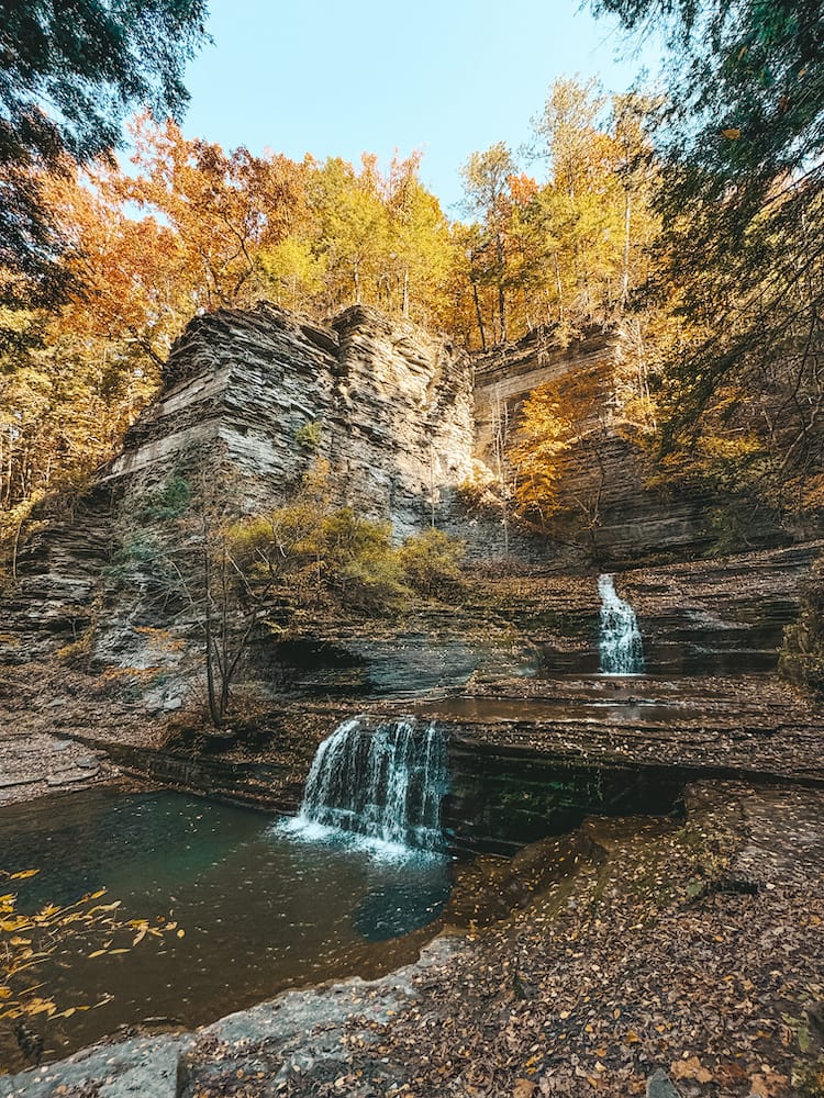 Several small waterfalls cascading down rocks inside a gorge in Buttermilk Falls State Park with yellow and orange fall leaves in the background.