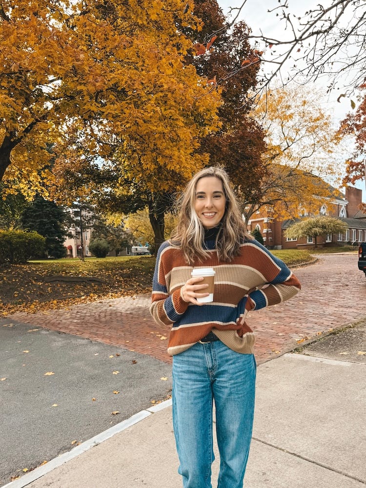 A woman in a striped sweater and denim jeans holding a cup of hot coffee standing in front of fall trees and a red brick road.