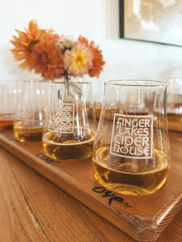 A tasting flight of hard cider from Finger Lakes Cider House on a wooden table with mums in a vase in the background.