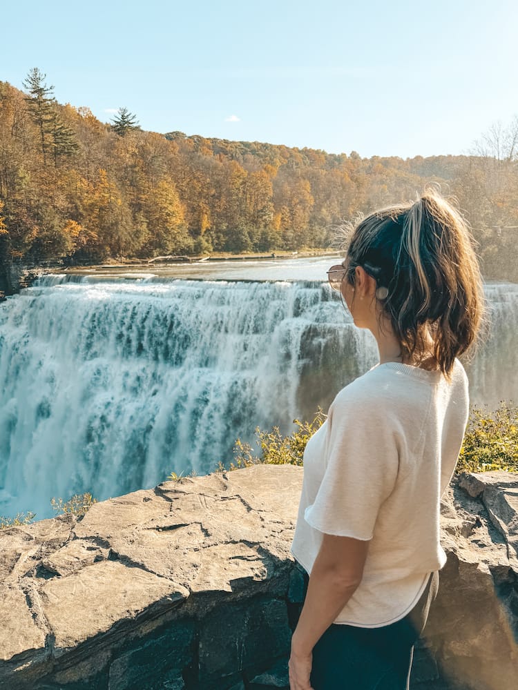A woman in a white shirt standing and looking at a waterfall with fall leaves in the background