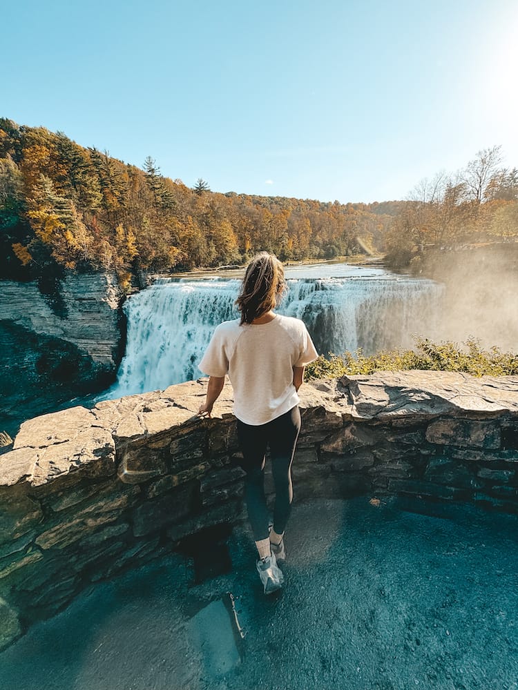 A woman in a white shirt and black leggings standing and overlooking a waterfall with fall trees in the background.
