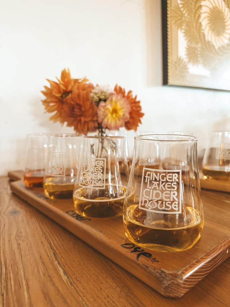 A flight of four hard ciders fron Finger Lakes Cider House, with a vase of orange and pink mums in the background.