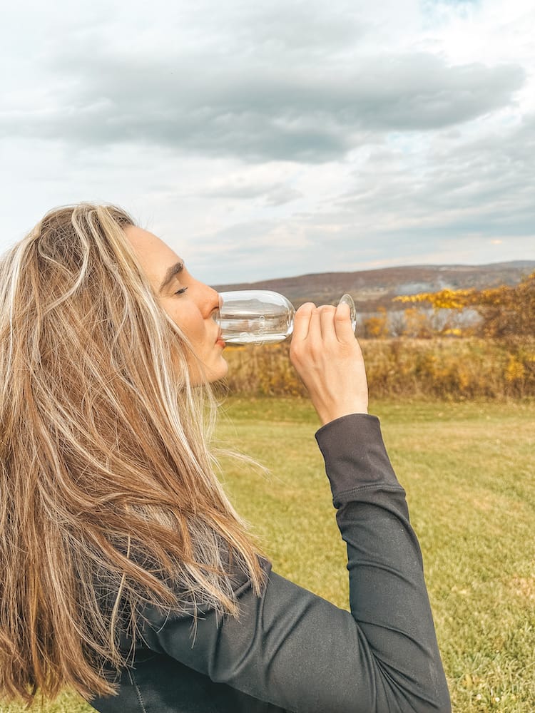 A woman with long brown hair wearing a black long-sleeve shirt, drinking a glass of wine in a vineyard in the Finger Lakes.