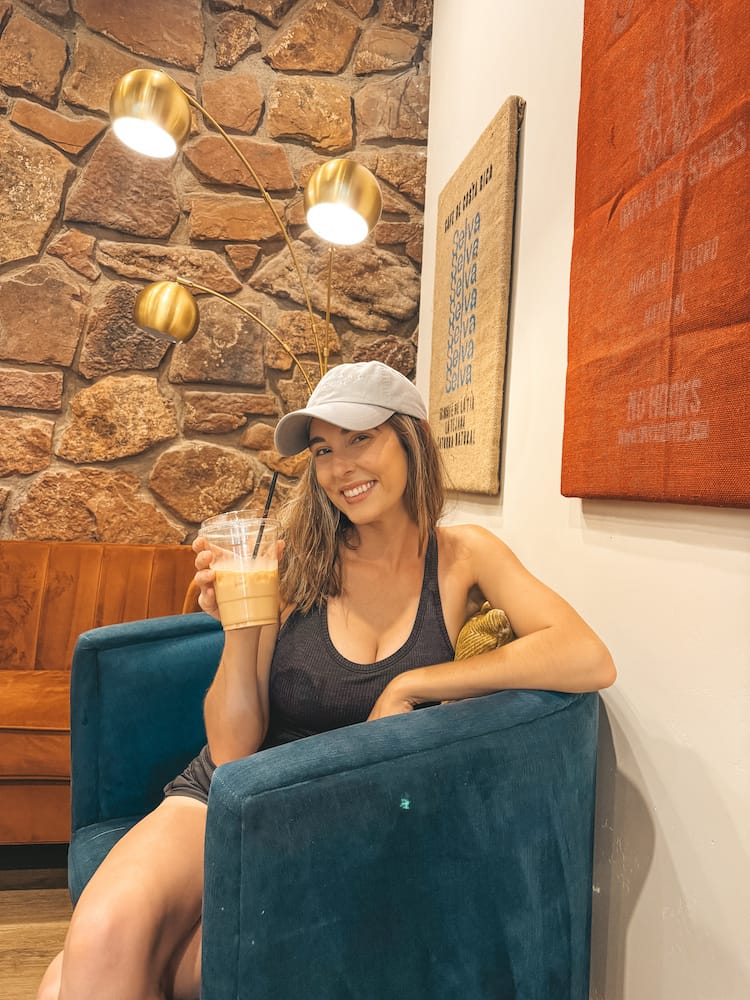 A woman sitting in a chair wearing a black tank top and gray cap in a coffee shop in Sedona holding an iced coffee in front of a stone wall.