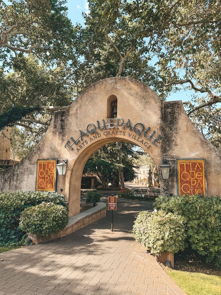 The entrance of Tlaquepaque, a shopping district in Sedona, surrounded by lush trees and bushes.