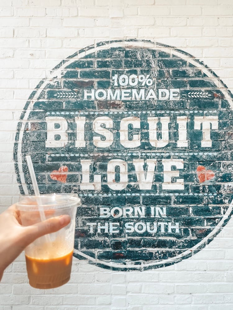 A half-finished iced coffee held in front of the black, white, and red sign for Biscuit Love painted on a white brick wall that reads" "100% Homemade Born in the South"