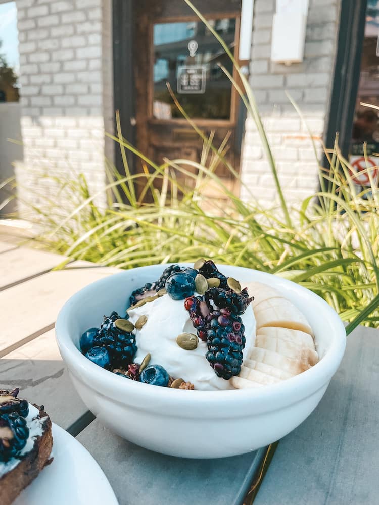 A yogurt parfait with bananas, blackberries, pumpkin seeds, and pomegranates sitting on a wooden table in front of greenery and a brick wall.