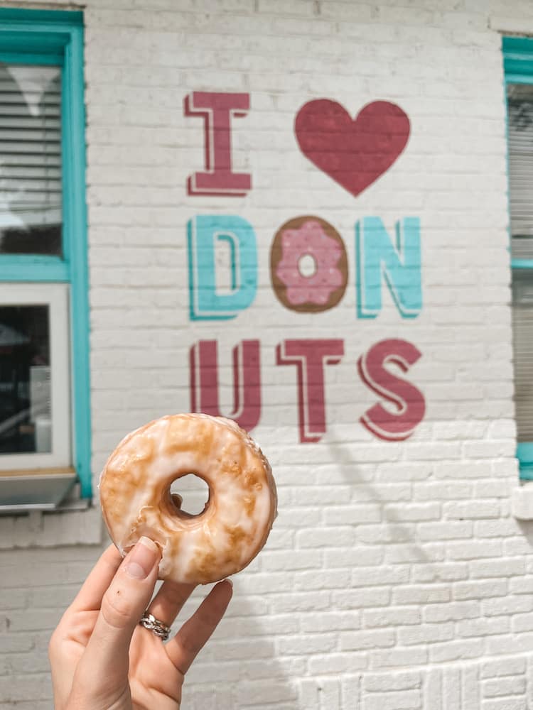 A donut being held in front of the "I Heart Donuts" sign painted on the side of the white brick building of the Five Daughters Bakery at 12 South.