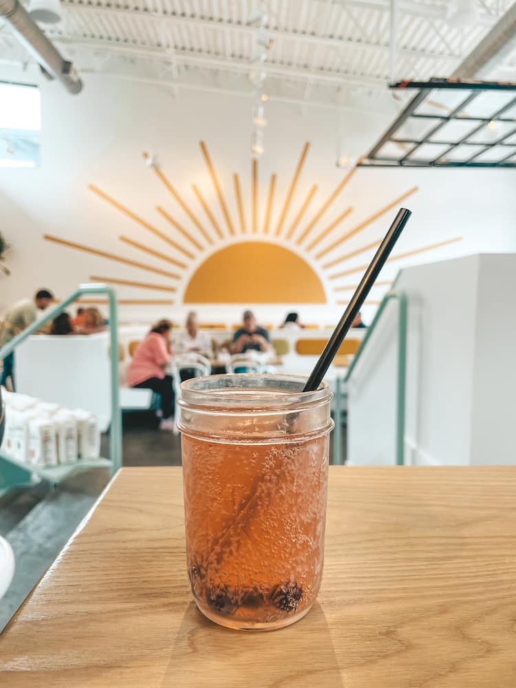 A mimosa in a mason jar with a black straw sitting on a wooden table inside a restaurant with a bright yellow sun painted on the white wall behind the drink.