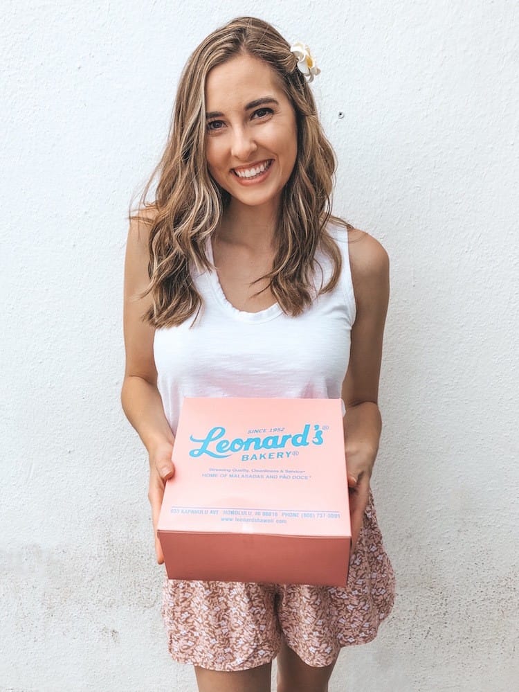 A woman with wavy brown hair in a white tank top and pink shorts holding a pink box that says "Leonard's Bakery" in blue letters