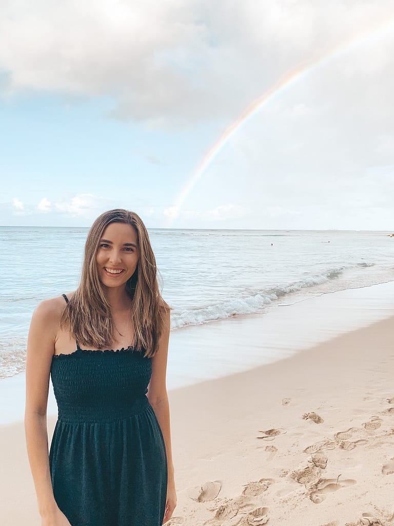 A girl with brown hair wearing a black jumpsuit standing in front of a rainbow on a beach.