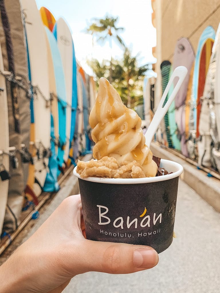A banana soft serve in a takeout container from Banan in front of several surfboards lining the way to Waikiki Beach.
