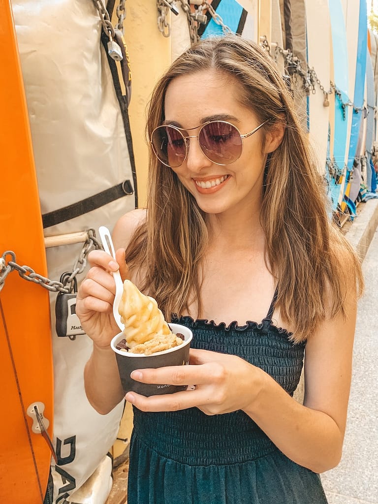A woman wearing sunglasses and a black jumpsuit with straight brown hair looks down at a banana soft serve ice cream in a black container before taking a bite.