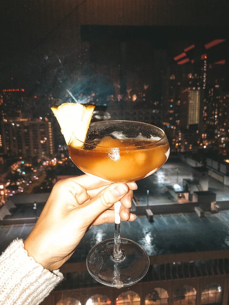 A frozen cocktail is being held in front of a panoramic view of Honolulu and Waikiki at night.