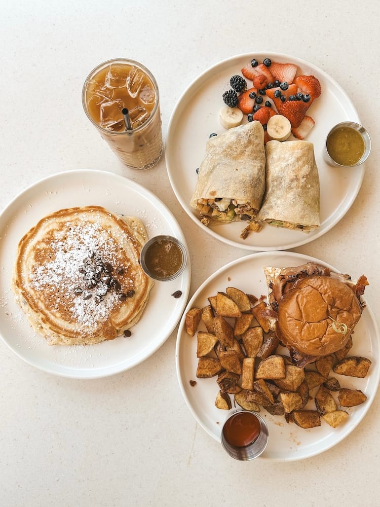 A brunch spread on a white table with an iced coffee, a pancake, breakfast burrito and fruit, and a breakfast sandwich with potatoes from The Eleanor in Old Town Scottsdale.