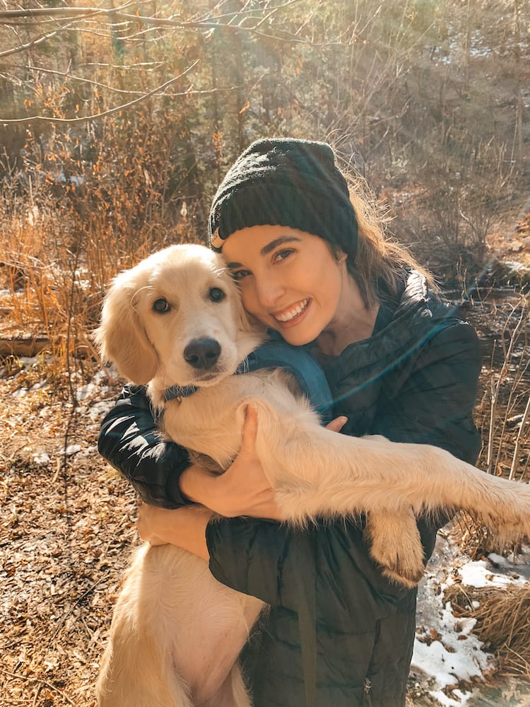 A woman wearing a black beanie and puffer jacket holding a golden retriever puppy and smiling at the camera