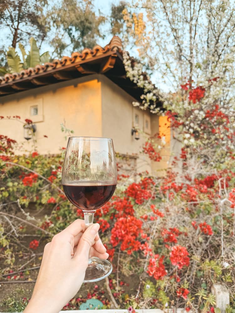 A glass of red wine being held in front of a Spanish-style building with bougainvilleas in front of it.