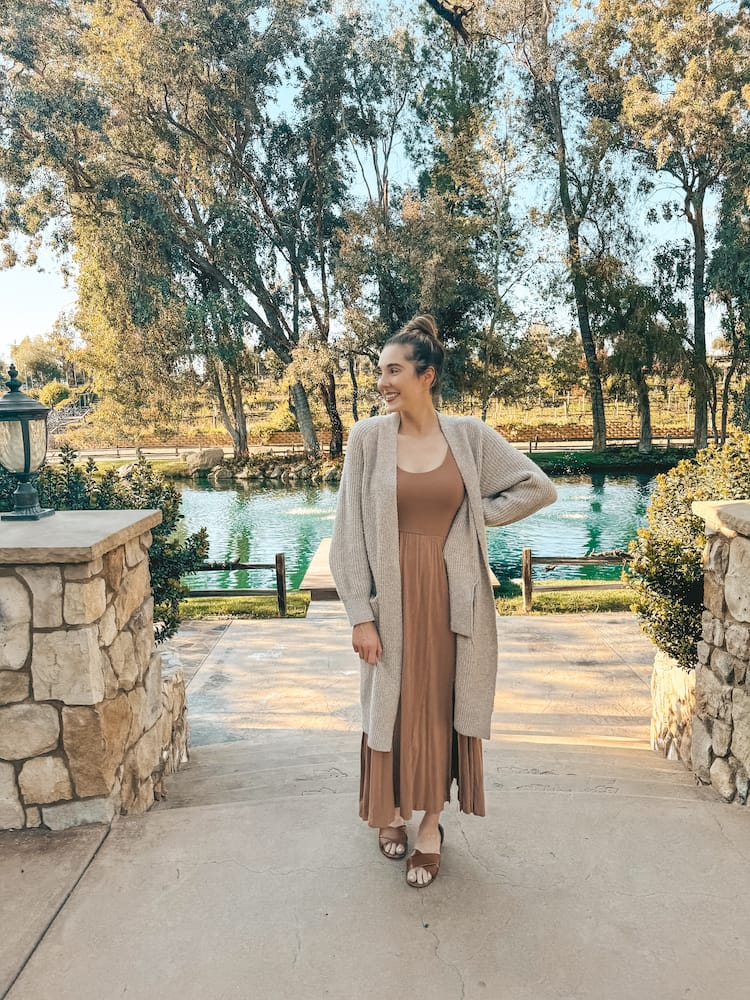 A woman wearing a brown dress and sweater standing in front of a pond at one of the best wineries in Temecula