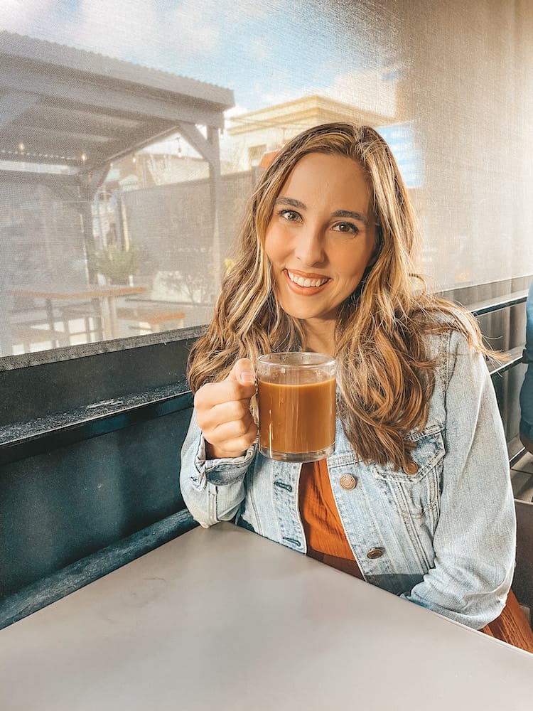 A woman holding a glass cup of coffee wearing a denim jacket and orange dress and sitting at a table.
