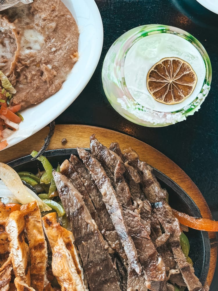 A spread of steak, chicken, and shrimp fajitas at a Mexican restaurant with a margarita and a plate of refried beans and rice.