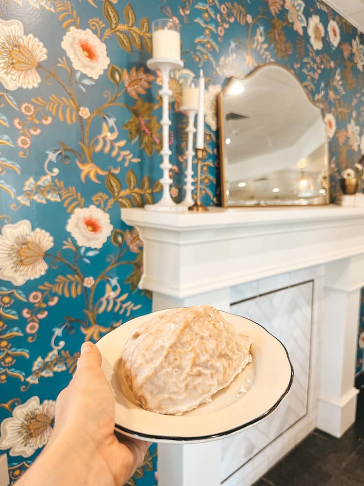 A glazed scone on a plate in front a blue floral wallpapered wall and a white fireplace.