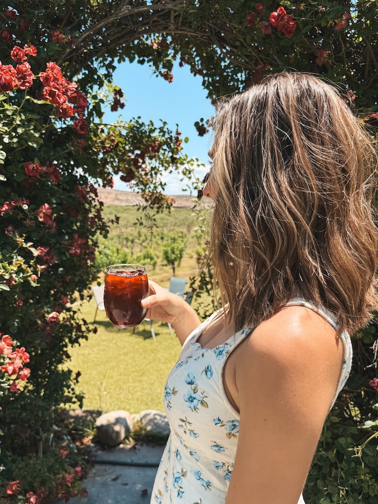 A woman in a white floral dress with short brown hair holding a glass on red wine in front of a greenery arch at a winery