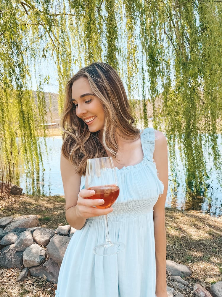 A woman in a blue sundress holding a glass of red wine in front of a lake with a green tree cascading in the background.