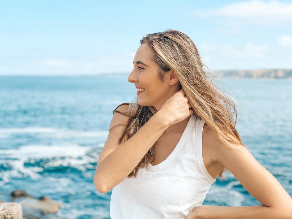 La Jolla or Coronado: A girl in a white shirt pushing her brown hear behind her shoulders in front of a blue ocean.