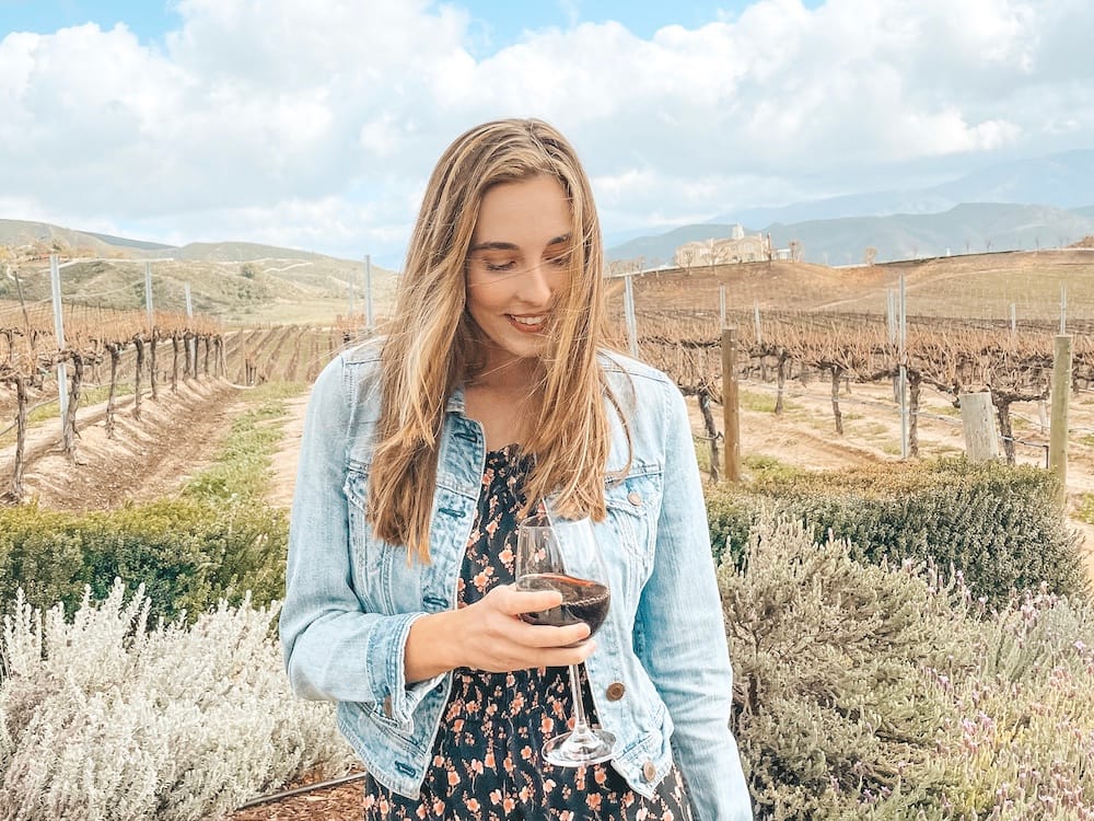 A woman standing in front of a vineyard with a glass of red wine.