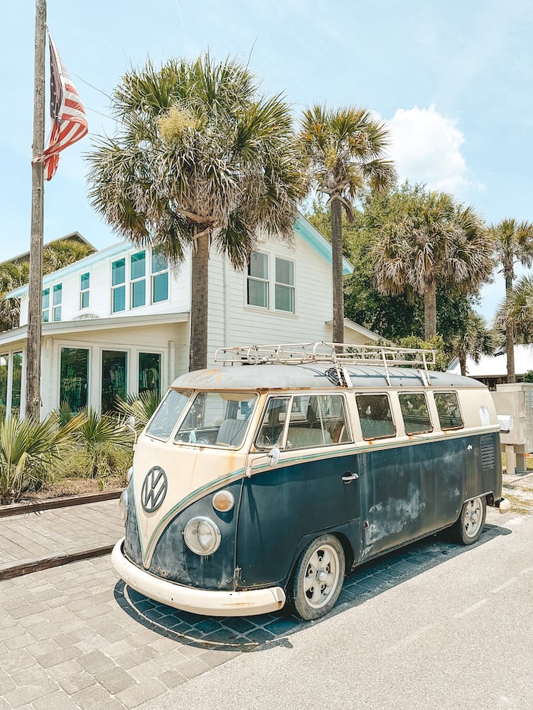 An old black and white VMW bus outside a building with palm trees and American flag in the background.