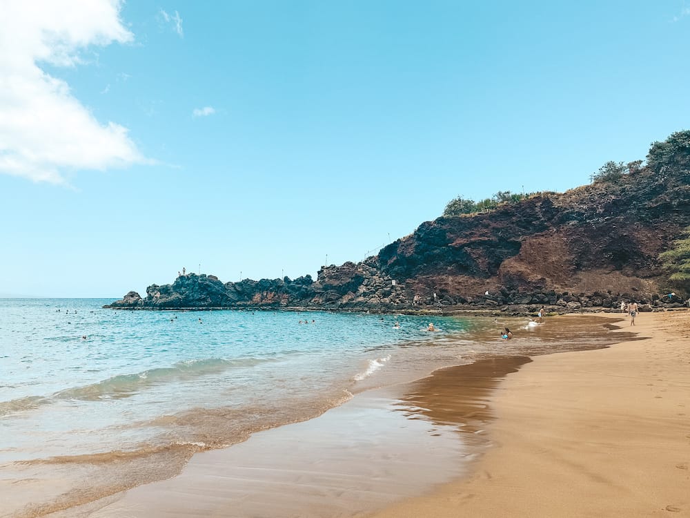A black rock sitting on a golden sand beach and jutting out into the water at Black Rock Beach, one of the best snorkeling spots in Maui.