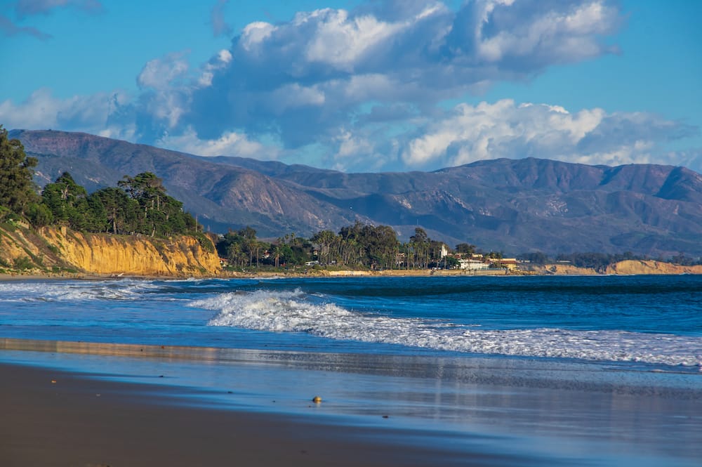 Bright blue waves rolling onto a dark sand beach with gorgeous hills in the background and cliffs covered in trees.