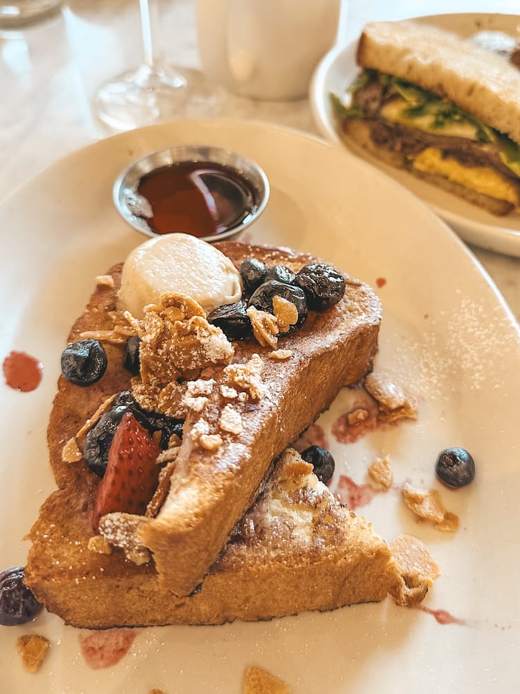 A stack of French toast with maple syrup, butter, fruit, and candied nuts on a white plate on a table with a sandwich in the background at Prep & Pastry in Old Town Scottsdale