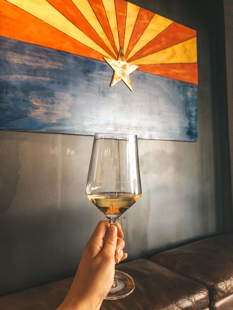 A glass of white wine held in front of the Arizona flag – featuring a blue bottom half and a yellow and red top half with a copper star in the center – in Old Town Scottsdale