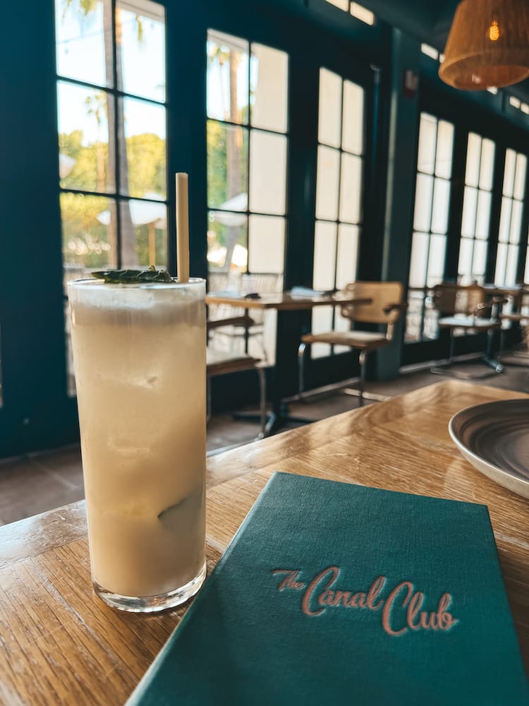 A pineapple and coconut cocktail sitting next to a green menu that says "Canal Club" in pink letters with windows in the background 