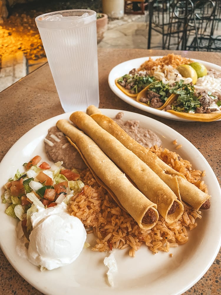 A white plate of Mexican food, with flautas, rice, refried beans, salsa, and sour cream, sits on a table with a plate of carne asada tacos and a cup of water.