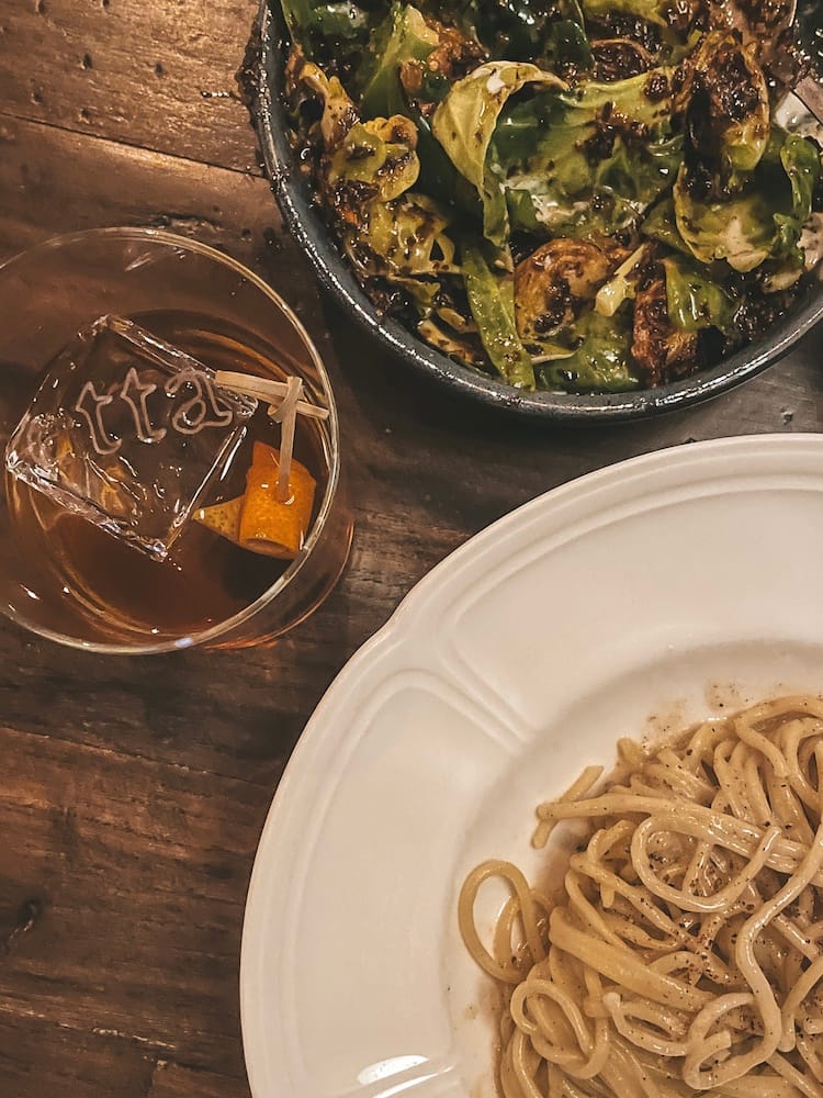 A wooden table with a bowl of Brussels sprouts, a plate of pasta, and a cocktail with ice branded with "Etta" at one of the best restaurants in Scottsdale at Scottsdale Quarter.