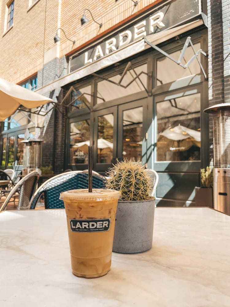 An iced coffee in a plastic cup sitting on a table in front of a small potted cactus. The background features a sign for the coffee shop, called Larder, as well as the entrance to the coffee shop and tan umbrellas.