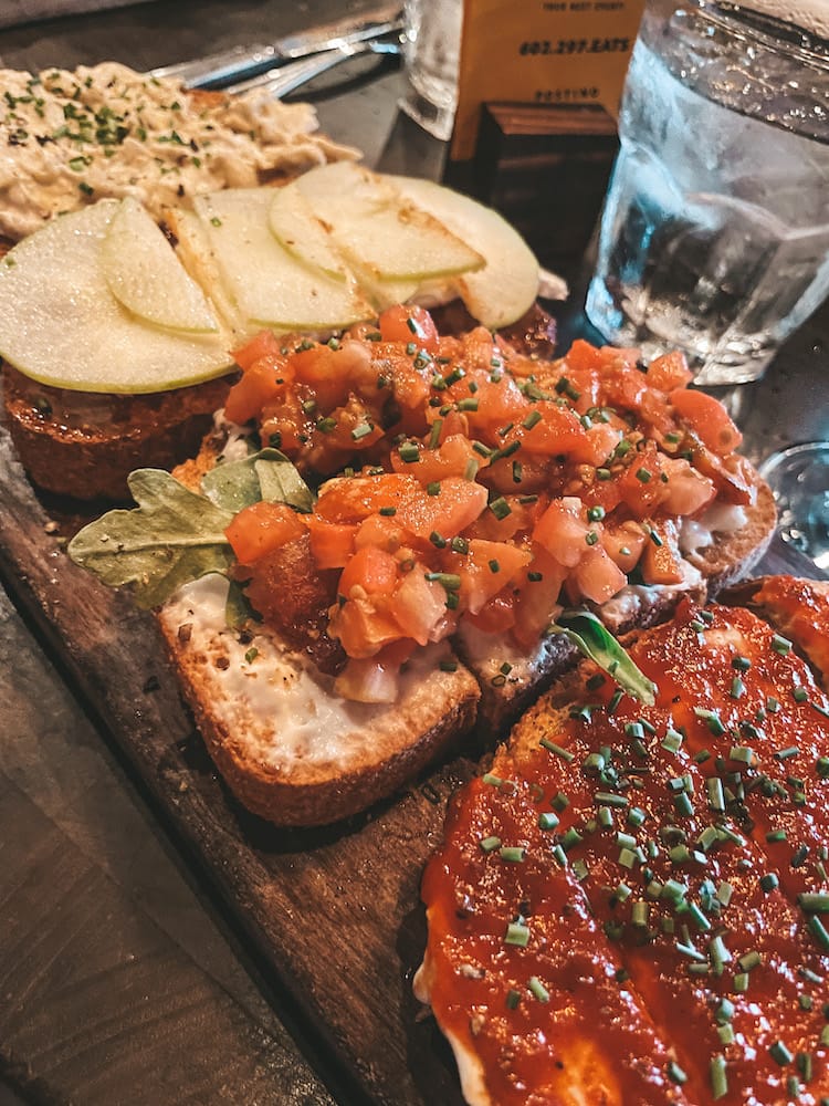 A wooden board with four bruschettas on it, one with chicken, one with apples, one with tomatoes, and one with jam.